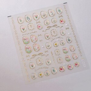 May include: Clear sheet of nail art stickers featuring various floral designs in pink, white, and yellow, with gold accents. The stickers include oval shapes, bows, and decorative frames. The text "just a little you" is also present.