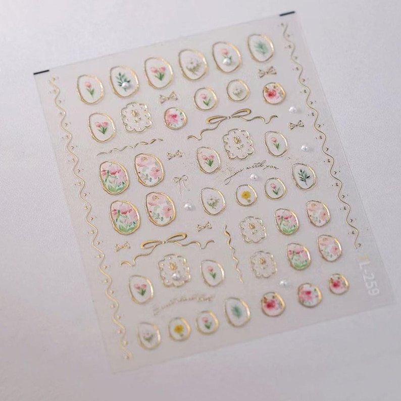May include: Clear sheet of nail art stickers featuring various floral designs in pink, white, and yellow, with gold accents. The stickers include oval shapes, bows, and decorative frames. The text "just a little you" is also present.
