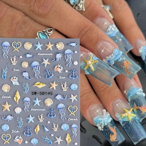 Puede incluir: Una hoja de pegatinas para uñas con una variedad de diseños con temática marina, incluyendo conchas, estrellas de mar, medusas, anclas y coral. La hoja está etiquetada como "SW-5D145" y presenta las palabras "Mar", "Coral" y "Gipppocampi".