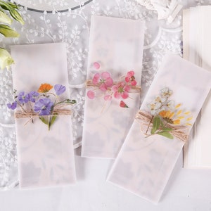 May include: Three white envelopes with pressed flowers and twine. The flowers are pink, purple, and yellow.