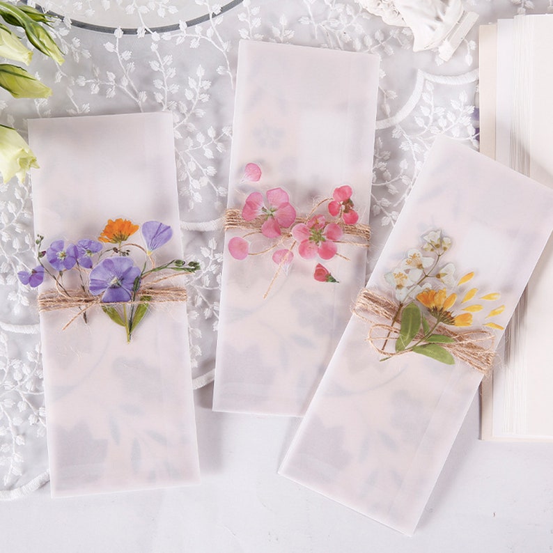 May include: Three white envelopes with pressed flowers and twine. The flowers are pink, purple, and yellow.