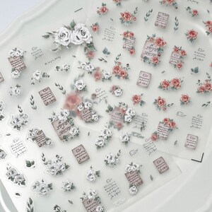 Puede incluir: Hojas transparentes de pegatinas decorativas con rosas blancas y rojas, hojas verdes y peque&ntilde;as notas de papel marr&oacute;n con texto. Las pegatinas tienen una est&eacute;tica vintage y rom&aacute;ntica, ideal para scrapbooking o manualidades.