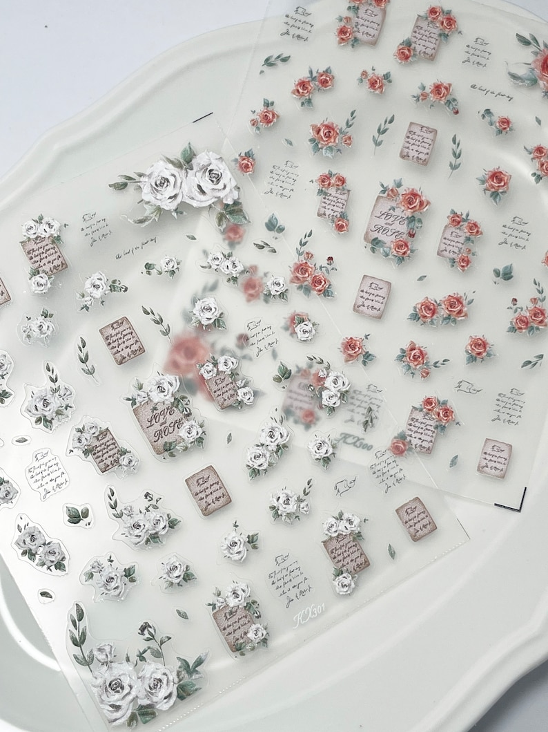 Puede incluir: Hojas transparentes de pegatinas decorativas con rosas blancas y rojas, hojas verdes y peque&ntilde;as notas de papel marr&oacute;n con texto. Las pegatinas tienen una est&eacute;tica vintage y rom&aacute;ntica, ideal para scrapbooking o manualidades.