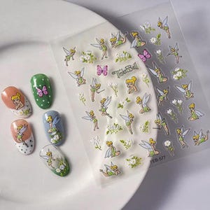 May include: A sheet of adhesive nail decals featuring Tinker Bell illustrations, small white flowers, and pink butterflies, alongside five oval-shaped nail tips decorated with the same Tinker Bell and floral designs, with the text TinkerBell and FEB-577.