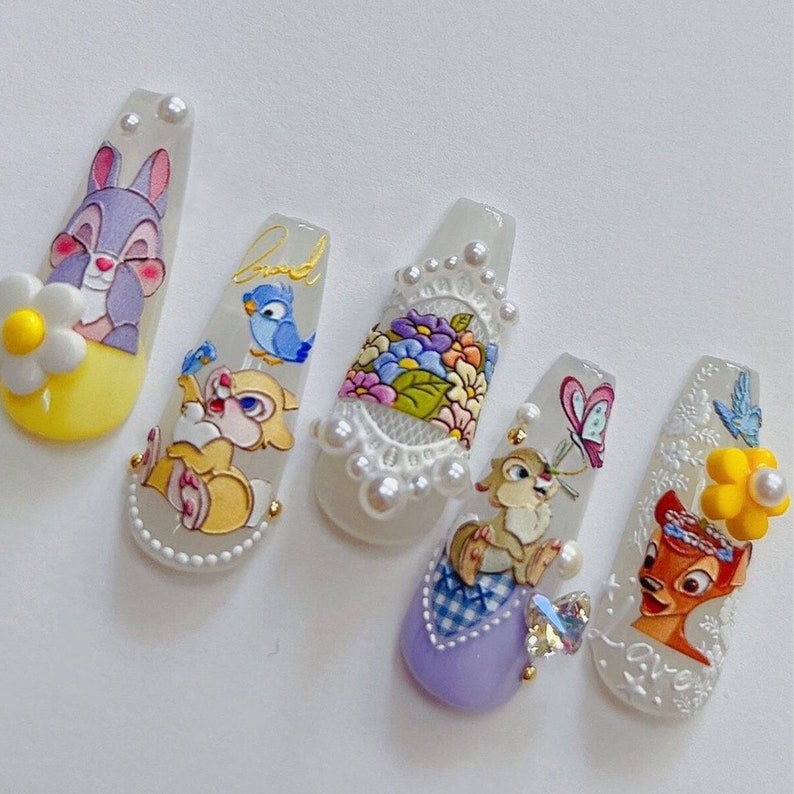 Peut inclure: Six pointes d'ongles blanches avec diff&eacute;rents motifs de personnages Disney. Les motifs comprennent un lapin, un oiseau, des fleurs, un papillon, un &eacute;cureuil et un cerf. Les pointes sont d&eacute;cor&eacute;es de perles et de paillettes.
