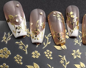 May include: Artificial nails with a gradient effect, transitioning from a light brown to white. The nails are adorned with gold floral designs and small rhinestones. Additional gold floral stickers are scattered around the nails.