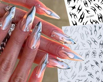 May include: Long, pointed fingernails with a pearlescent finish and silver flame designs. Each nail is adorned with small, sparkling rhinestones. Two sheets of nail art stickers with black tribal designs are visible.