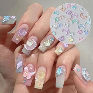 Puede incluir: Un conjunto de dijes iridiscentes para uñas con formas de mariposas, estrellas y lunas crecientes en tonos pastel de rosa, azul, amarillo y morado. Estos pequeños adornos translúcidos y brillantes se muestran aplicados en uñas manicuradas.