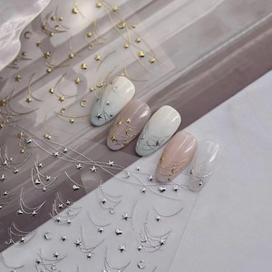 May include: A sheet of adhesive nail decals featuring metallic gold and silver designs of crescent moons, stars, and delicate hanging chains. Five artificial nail tips are displayed, each decorated with these celestial stickers in various patterns.