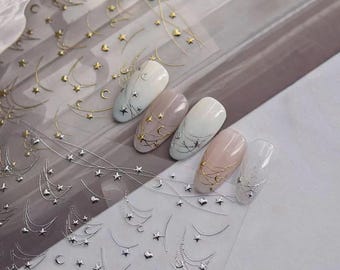 May include: A sheet of adhesive nail decals featuring metallic gold and silver designs of crescent moons, stars, and delicate hanging chains. Five artificial nail tips are displayed, each decorated with these celestial stickers in various patterns.