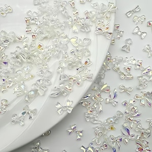 May include: A collection of clear, iridescent bow-shaped nail art decorations scattered on a white surface. The bows vary in size and reflect light, creating a shimmering effect. These decorative bows are ideal for nail art.