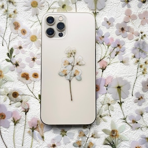 May include: A white iPhone with a pressed flower design on the back. The phone has a white background and a black camera lens.
