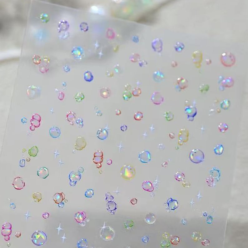 May include: Clear sticker sheet with iridescent bubble and star designs. The bubbles are in various sizes and colors, including pink, blue, green, and yellow. The stars are small and white.