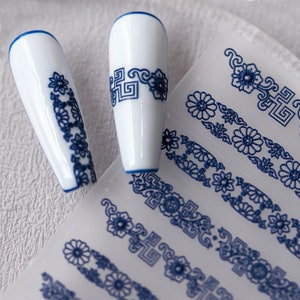 May include: Blue and white nail art stickers featuring a floral and geometric design. The stickers are on a clear sheet and are ready to be applied to nails.