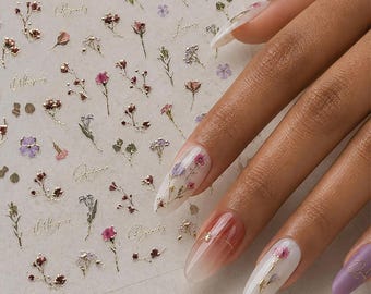 May include: A sheet of adhesive nail decals featuring small metallic gold floral illustrations, delicate pink and purple flower accents, and cursive script text. The designs are applied to fingernails with varying base colors and finishes.