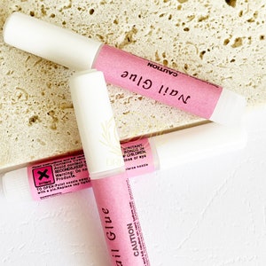 May include: Three tubes of nail glue with white caps and pink labels. The labels read "Nail Glue" and "CAUTION" in black text. The background is a textured, light-colored surface.