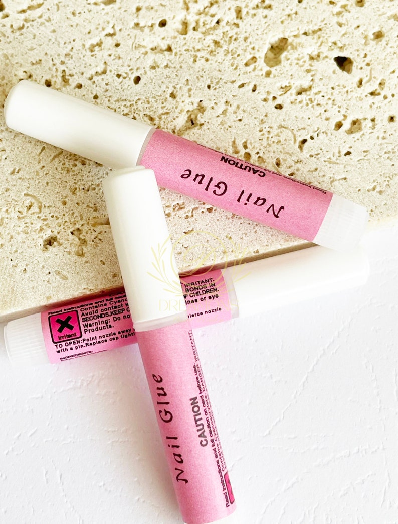 May include: Three tubes of nail glue with white caps and pink labels. The labels read "Nail Glue" and "CAUTION" in black text. The background is a textured, light-colored surface.