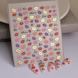 Pu&ograve; includere: Foglio di adesivi per nail art con vari disegni di labbra colorate in rosso, rosa, viola e oro. Il foglio include parole come "Kiss", "Love" ed "Enthusiasm". Diverse unghie decorate sono esposte sotto.