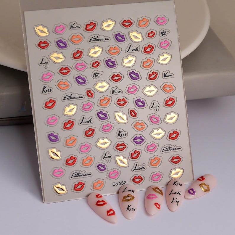 Pu&ograve; includere: Foglio di adesivi per nail art con vari disegni di labbra colorate in rosso, rosa, viola e oro. Il foglio include parole come "Kiss", "Love" ed "Enthusiasm". Diverse unghie decorate sono esposte sotto.