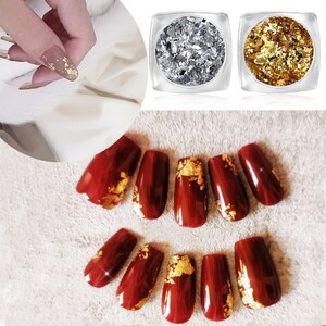 May include: Two small jars of gold and silver nail flakes. The jars are clear plastic with a silver lid. The flakes are loose and scattered inside the jars. A set of ten red press-on nails with gold flakes on top.