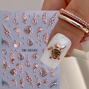 May include: Rose gold nail art stickers featuring various sea creatures and nautical elements, including seahorses, anchors, and fish. The stickers are on a clear sheet with the text "SW-5D180". A nail with a gold turtle charm is also visible.