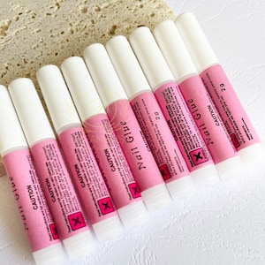 May include: A collection of nail glue tubes. Each tube has a white cap and a pink label with black text, including the word "CAUTION" and a warning symbol. The tubes are arranged in a row, ready for use.