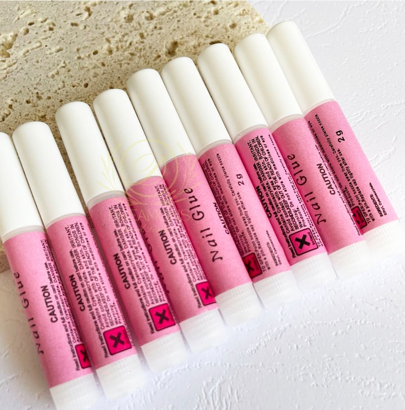 May include: A collection of nail glue tubes. Each tube has a white cap and a pink label with black text, including the word "CAUTION" and a warning symbol. The tubes are arranged in a row, ready for use.