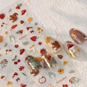 May include: A sheet of adhesive nail decals featuring autumn-themed illustrations including squirrels, red and white mushrooms, colorful leaves, berries, and flowers. The sheet includes the text Autumn in a cursive font and four decorated artificial nails.