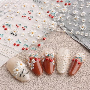 May include: Nail art stickers with white daisies and red cherries. The stickers are on a clear sheet and are designed to be applied to nails. The stickers are shown on a set of five nails with a brown base coat.