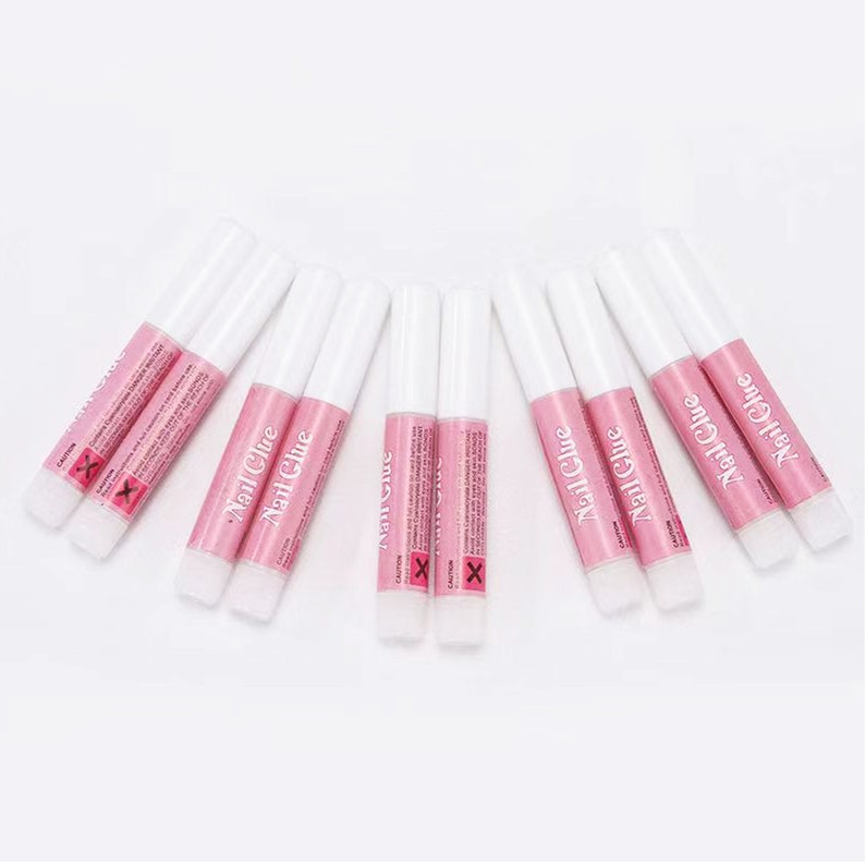 May include: A collection of nail glue tubes with white caps and pink labels. The labels read "Nail Glue" in a cursive font. The tubes are arranged in a fan shape against a white background. The word "CAUTION" and an "X" symbol are also visible.
