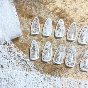 May include: A selection of white lace nail art designs displayed on a gold surface. The image features clear nail tips adorned with detailed lace patterns, and a roll of similar lace material. The designs incorporate floral and geometric patterns, ideal for a delicate manicure.