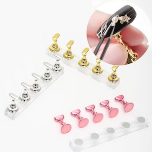 以下が含まれることがあります： 透明なアクリルスタンドが付いたネイルアートツールのセット。ツールはゴールド、シルバー、ピンクの色です。ツールは、ネイルポリッシュやデコレーションを塗布する際に、つけ爪を固定するために使用されます。