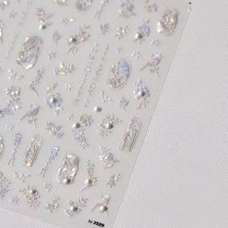 Puede incluir: L&aacute;mina transparente de pegatinas iridiscentes para decoraci&oacute;n de u&ntilde;as. Las pegatinas presentan varios dise&ntilde;os, incluyendo flores, p&aacute;jaros y formas abstractas. Las pegatinas tienen un efecto 3D y est&aacute;n adornadas con peque&ntilde;os detalles perlados. La esquina inferior derecha de la l&aacute;mina tiene el texto "to 3529".
