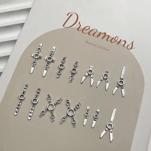 May include: An assortment of silver metal charms in diverse shapes, such as crosses, circles, and compass designs. The charms are displayed on a light-toned surface, with the words "Dreamons" and "Based London" printed in a script font.