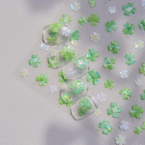 May include: Clear nail art stickers featuring green shamrocks and white flowers. The stickers are arranged on a clear sheet, with some applied to clear nail tips. The design is perfect for St. Patrick's Day or spring.