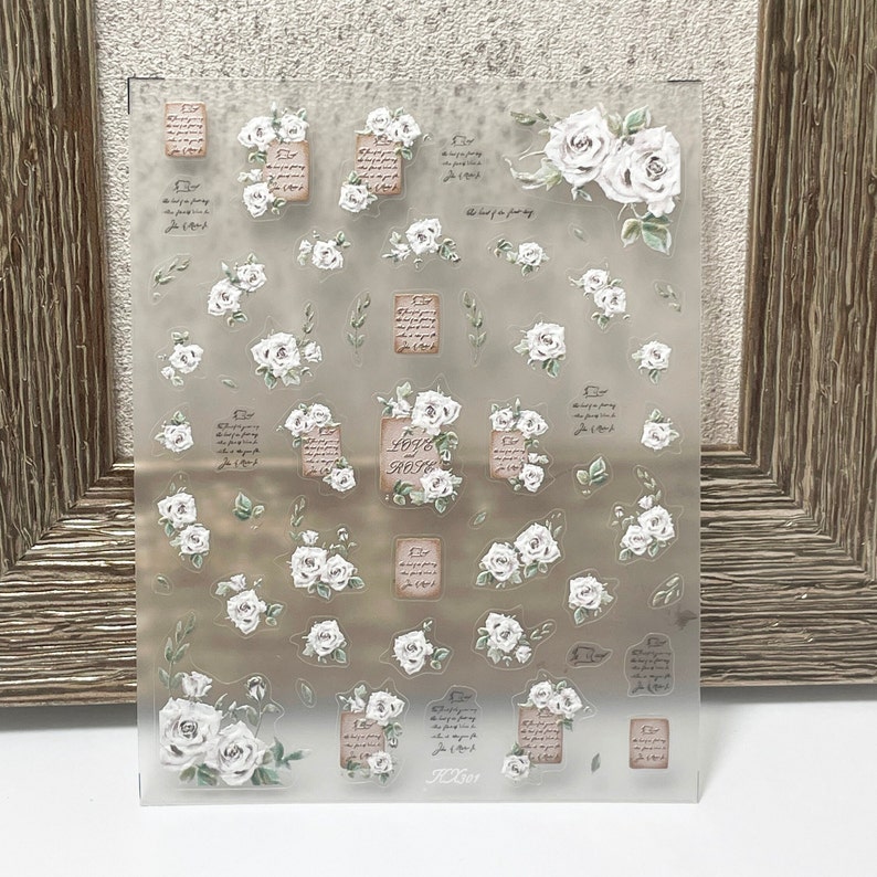 Puede incluir: Hoja de pegatinas transparente con ilustraciones de rosas blancas y texto. La hoja incluye rosas de varios tama&ntilde;os, ramitas y dise&ntilde;os rectangulares con escritura y las palabras "LOVE and ROSE".