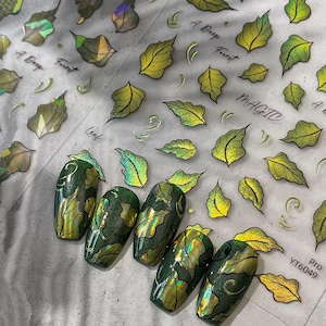 May include: Green and gold nail art with leaf designs. The nails are painted with a dark green base and adorned with gold leaf accents. The image also shows leaf-shaped nail art stickers and the text "A Deep Forest" and "Magic".