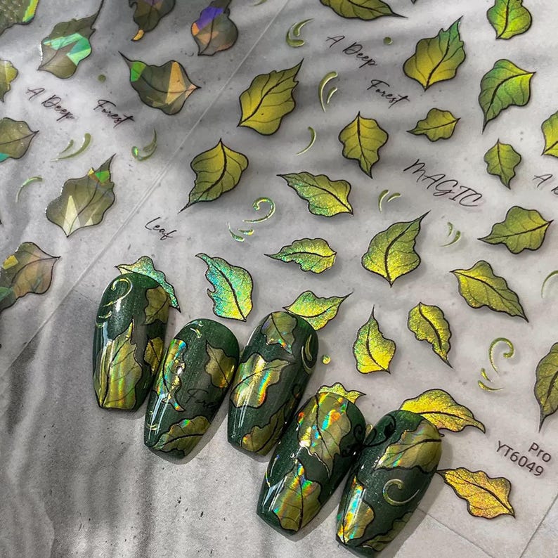 May include: Green and gold nail art with leaf designs. The nails are painted with a dark green base and adorned with gold leaf accents. The image also shows leaf-shaped nail art stickers and the text "A Deep Forest" and "Magic".