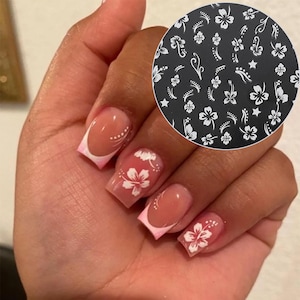 Könnte beinhalten: Hände mit manikürten Nägeln mit French-Tip-Design und rosa Akzent. Jeder Nagel ist mit einem weißen Hibiskusblüten-Design verziert. Eine kreisförmige Nagelkunstschablone mit weißen Blumenmustern ist ebenfalls sichtbar.