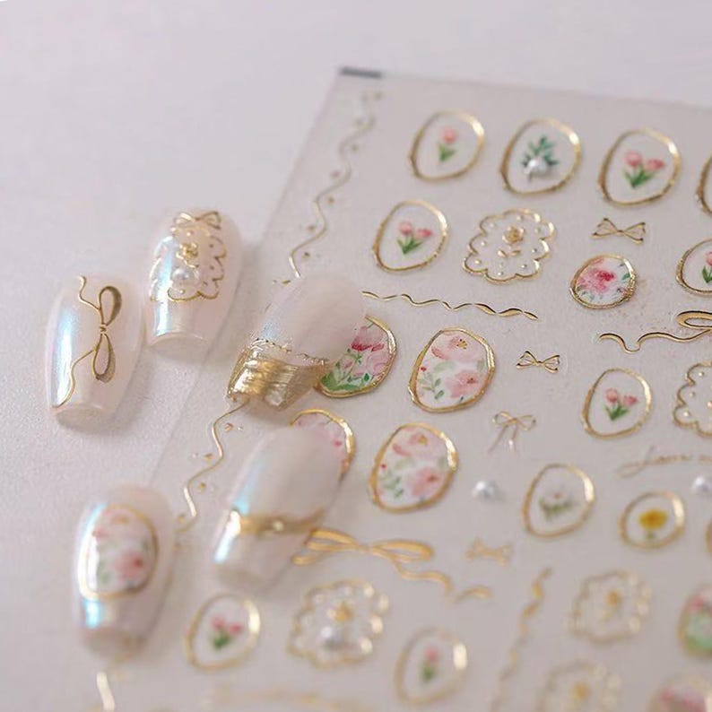 May include: Nail art stickers featuring floral designs and gold accents. The stickers include various shapes, such as oval frames, bows, and floral patterns. The nails are painted with a pearlescent polish, and the stickers are on a clear sheet.