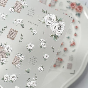 Puede incluir: L&aacute;mina transparente de pegatinas florales con rosas blancas, hojas verdes y escritura vintage. Las pegatinas incluyen rosas individuales, racimos de rosas y peque&ntilde;os ramos. Un plato blanco con calcoman&iacute;as de rosas en el fondo.