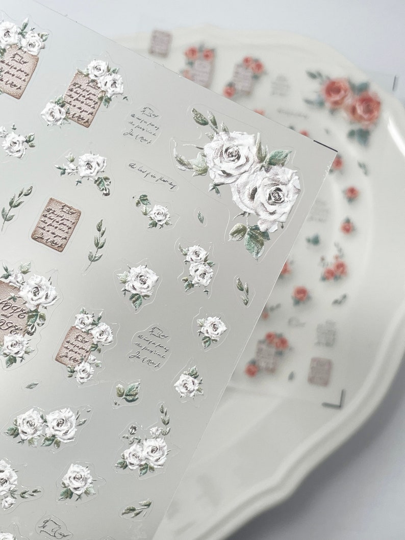 Puede incluir: L&aacute;mina transparente de pegatinas florales con rosas blancas, hojas verdes y escritura vintage. Las pegatinas incluyen rosas individuales, racimos de rosas y peque&ntilde;os ramos. Un plato blanco con calcoman&iacute;as de rosas en el fondo.