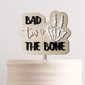 May include: A wooden cake topper with a skeleton hand making a peace sign and the text "Bad two the bone".