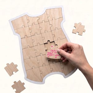 May include: A light-colored wooden jigsaw puzzle shaped like a baby onesie. The puzzle features a white border and multiple interlocking pieces. One piece is held in place, displaying the handwritten pink text: Can't wait to meet you.