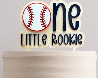 May include: A cake topper featuring a baseball graphic replacing the letter O in the word one, with the text LITTLE ROOKIE underneath. The design is layered with dark blue lettering on a light wood background, creating a dimensional effect.