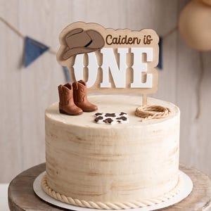 May include: A round cake with light brown textured frosting and a rope-style border. The cake topper is a wooden sign reading "Caiden is ONE" with a brown cowboy hat icon. Decorations include two brown cowboy boots, a lasso, and a cow-print piece.