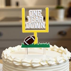 May include: A white frosted cake with a decorative topper that reads "ONE YEAR DOWN". The topper features a yellow goalpost, a brown football, and green grass. The cake is on a wooden table.