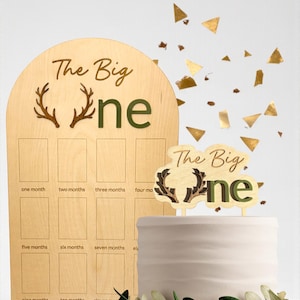 May include: Wooden milestone board and cake topper set. The board has spaces for monthly photos and reads "The Big One." The cake topper also says "The Big One." Both feature antlers and green lettering. Gold confetti is scattered around.