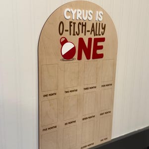 May include: An arched wooden milestone board featuring the text CYRUS IS O-FISH-ALLY ONE with a red and white fishing bobber graphic. The board includes twelve rectangular slots labeled from one month to one year for photo placement.