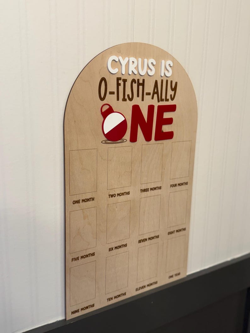 May include: An arched wooden milestone board featuring the text CYRUS IS O-FISH-ALLY ONE with a red and white fishing bobber graphic. The board includes twelve rectangular slots labeled from one month to one year for photo placement.